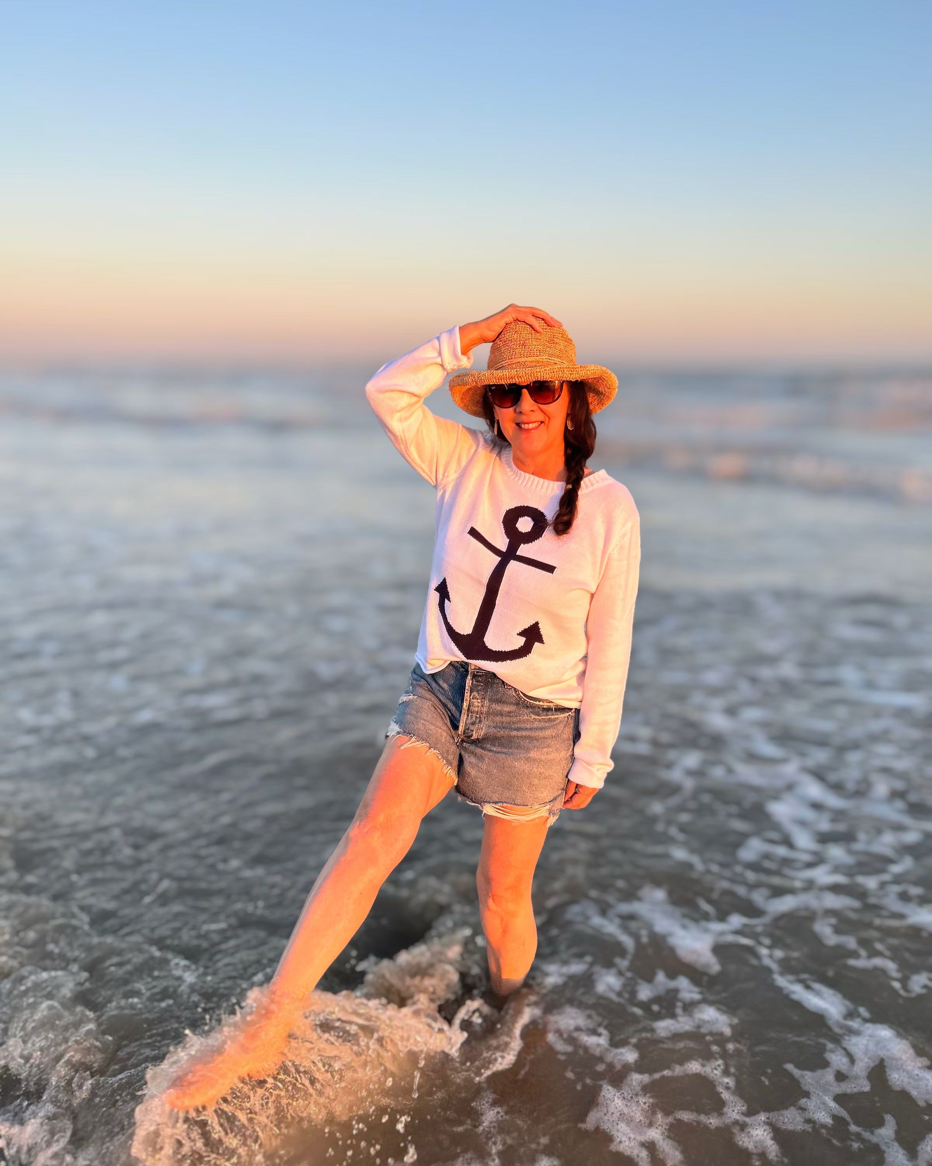 Person standing in shallow water at the beach wearing a hat and sunglasses.