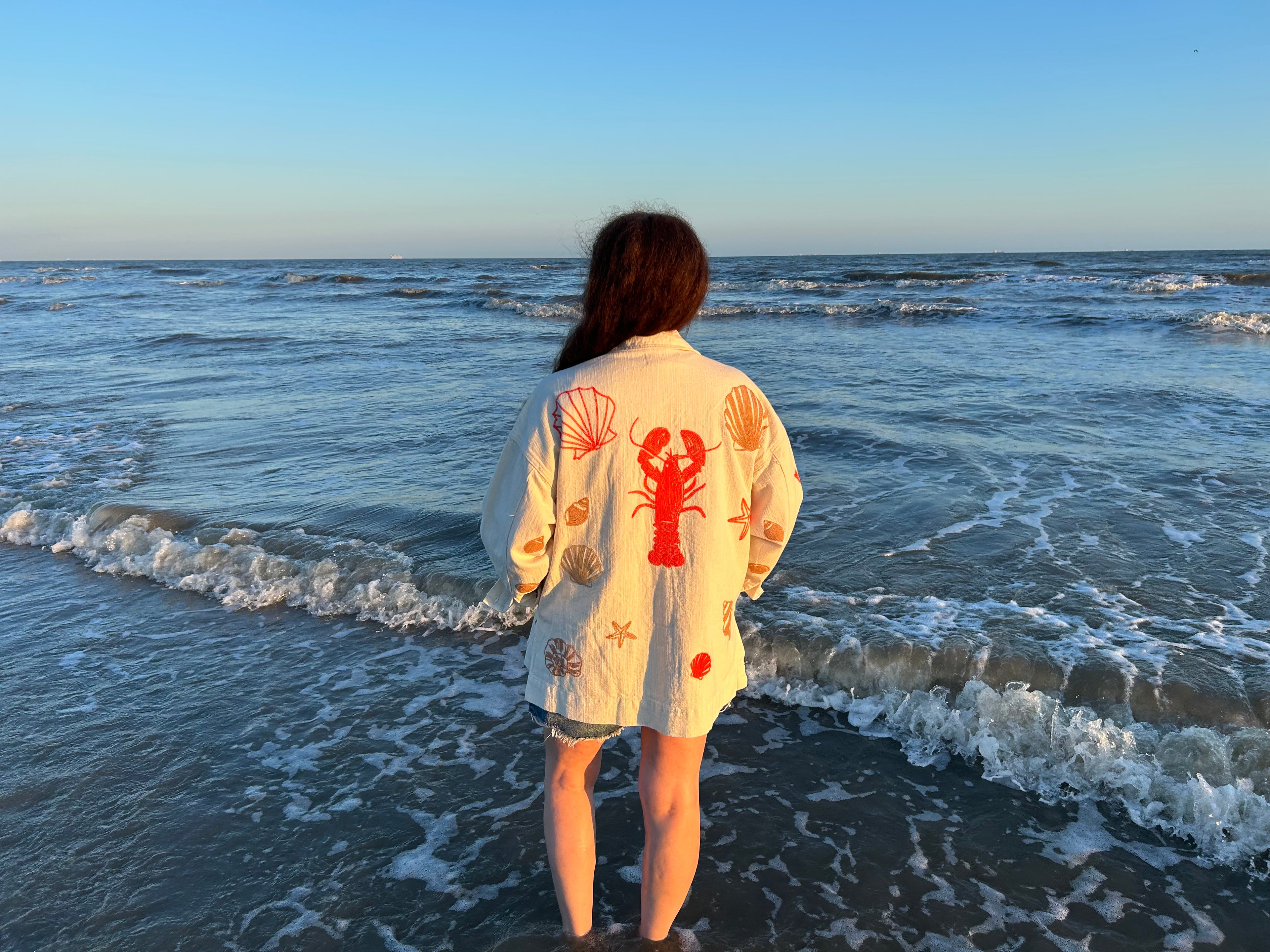 Person standing on a beach wearing a decorative shirt with shell and lobster designs.