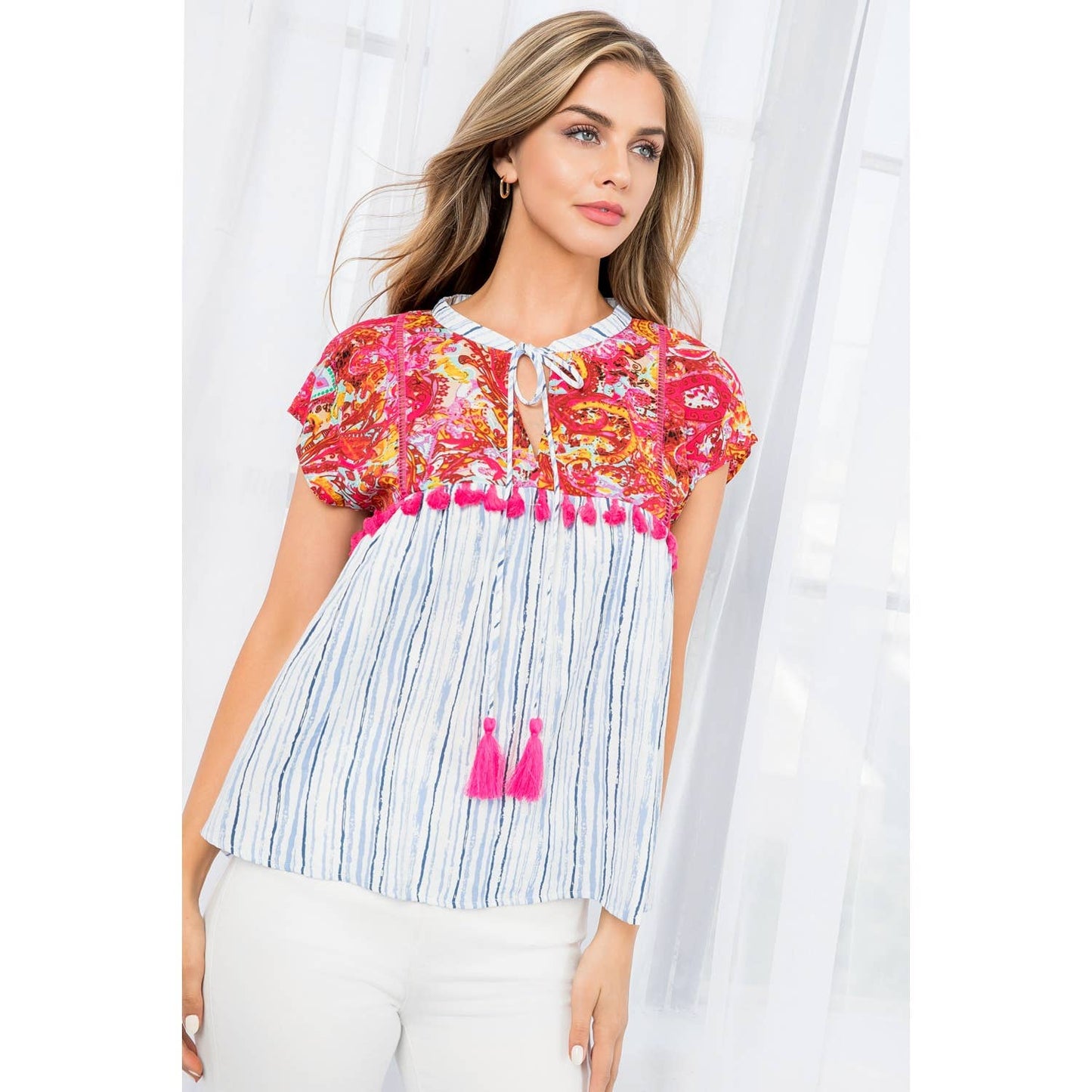 Woman wearing vibrant mixed print babydoll top with paisley and stripes, hot pink tassel details, and tie neckline